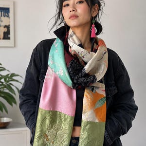 以下が含まれることがあります： ピンク、ミントグリーン、オリーブグリーン、オレンジ、黒の色合いの様々な布パネルを組み合わせたパッチワークスカーフ。スカーフには鶴、花柄、幾何学模様などの複雑なデザインが施されており、黒のデニムジャケットの上に着用されています。