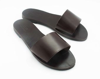 May include: A pair of brown leather slide sandals. The sandals have a simple design with a wide strap across the top of the foot. The soles are dark brown and the sandals appear to be new.