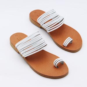 May include: A pair of white leather sandals with a toe loop and multiple straps across the top. The sandals have a brown leather sole and are displayed on a white background. The sandals are a size 36.