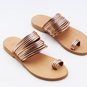 May include: A pair of rose gold sandals with a toe loop and multiple straps across the top. The sandals have a light brown leather footbed and a simple design, perfect for warm weather. The sandals are displayed on a white background.
