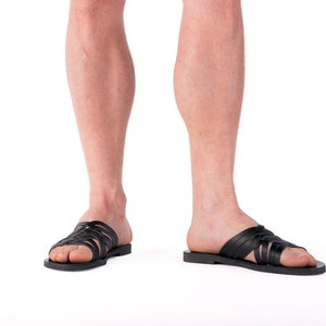 Puede incluir: Sandalias de cuero negro con un dise&ntilde;o de correas trenzadas. Las sandalias tienen suela plana y se llevan en los pies de una persona. La imagen est&aacute; sobre un fondo blanco.