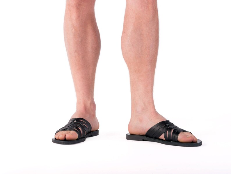 Puede incluir: Sandalias de cuero negro con un dise&ntilde;o de correas trenzadas. Las sandalias tienen suela plana y se llevan en los pies de una persona. La imagen est&aacute; sobre un fondo blanco.