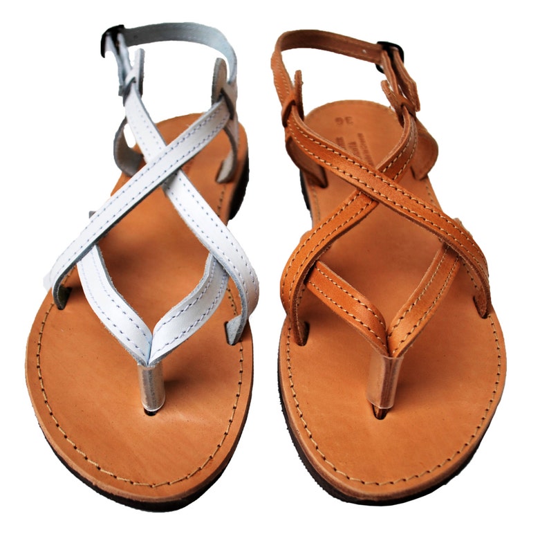 May include: Two pairs of leather sandals. One pair features white straps, the other brown. Both have a thong-style toe post and crisscross straps. The soles are light brown with dark brown edges. Adjustable ankle straps with buckles are visible.