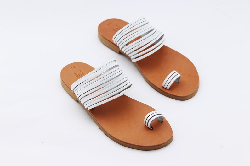 May include: A pair of white leather sandals with a toe loop and multiple straps across the top. The sandals have a brown leather sole and are displayed on a white background. The sandals are a size 36.