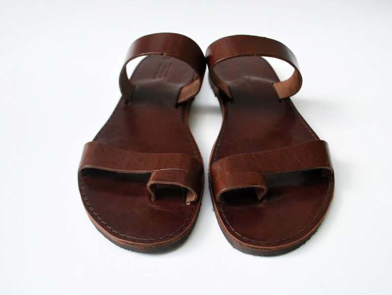 May include: A pair of brown leather sandals with a toe loop and ankle strap. The sandals have a simple, minimalist design, with visible stitching around the edges. The leather appears to be smooth and well-worn.