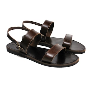 May include: A pair of brown leather sandals with a simple design. The sandals have a flat sole and two straps across the foot, with an ankle strap. The sandals are displayed on a white background.