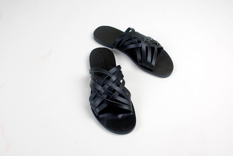 Puede incluir: Un par de sandalias de cuero negro con un dise&ntilde;o de correas trenzadas. Las sandalias se muestran sobre una superficie blanca. Tienen una suela plana y un estilo sencillo y minimalista, adecuado para uso casual.