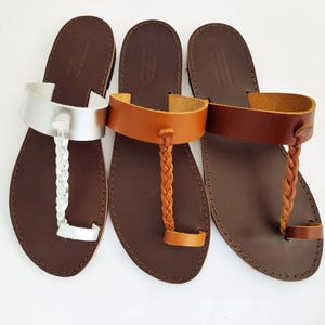 May include: Three leather sandals in a row. The sandals have brown soles and different colored straps. The first sandal has a silver strap and a braided toe strap. The second sandal has a tan strap and braided toe strap. The third sandal has a brown strap and braided toe strap.