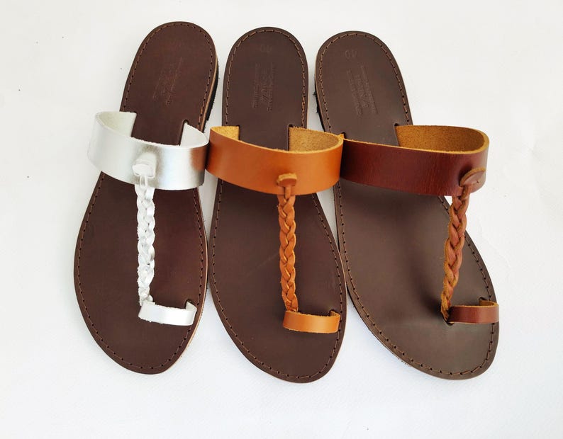 May include: Three leather sandals in a row. The sandals have brown soles and different colored straps. The first sandal has a silver strap and a braided toe strap. The second sandal has a tan strap and braided toe strap. The third sandal has a brown strap and braided toe strap.