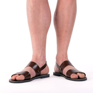 Op de afbeelding: Bruine leren sandalen met twee bandjes over de voet en een enkelbandje. De sandalen hebben een eenvoudig ontwerp met een donkerbruine kleur en een zwarte zool. De afbeelding toont de sandalen van voren.