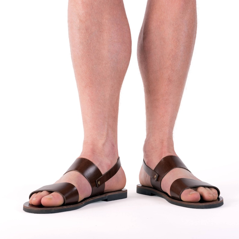 Op de afbeelding: Bruine leren sandalen met twee bandjes over de voet en een enkelbandje. De sandalen hebben een eenvoudig ontwerp met een donkerbruine kleur en een zwarte zool. De afbeelding toont de sandalen van voren.