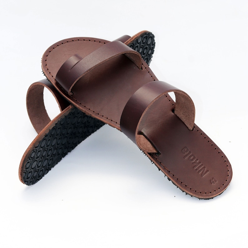 May include: Brown leather sandals with a toe loop and two straps. The sandals have a dark brown sole with a textured pattern. The word "Nikola" is embossed on the footbed.