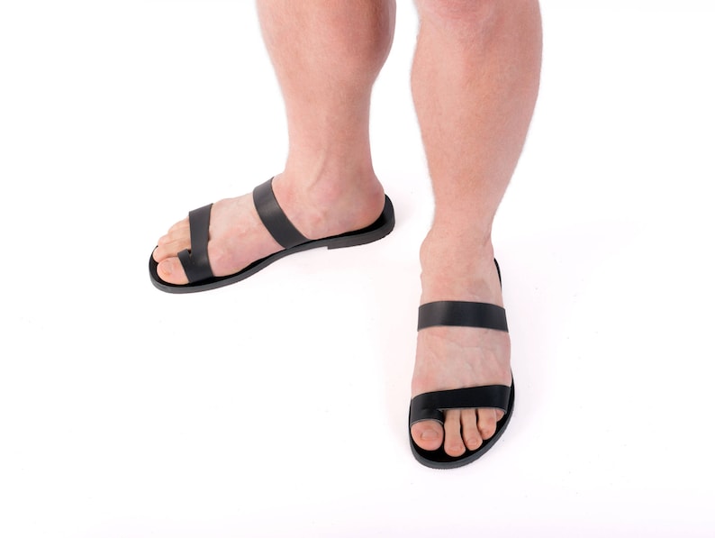 May include: Black leather sandals with a toe loop and two straps. The sandals are shown from a low angle, with the feet and lower legs of a person visible against a white background. The sandals appear to be flat and minimalist in design.