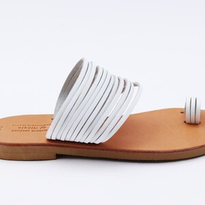 May include: White leather sandals with a toe loop and multiple straps across the top. The sandals have a light brown sole and are designed for a comfortable fit. The footwear is suitable for warm weather.