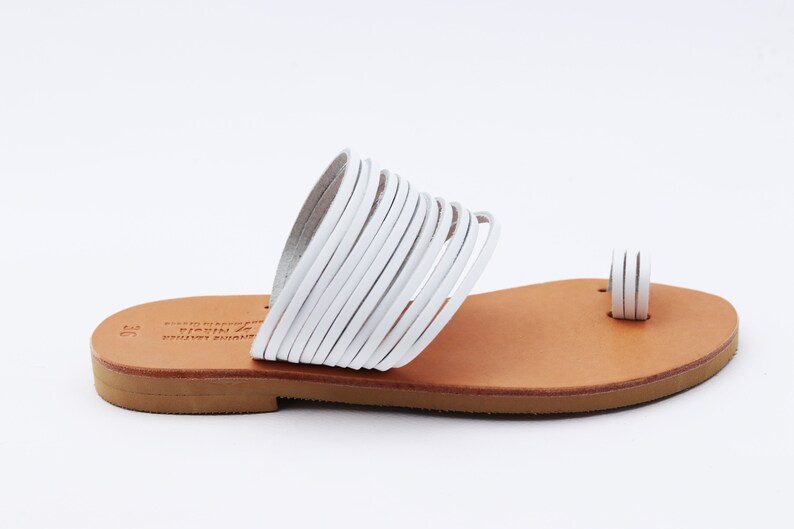May include: White leather sandals with a toe loop and multiple straps across the top. The sandals have a light brown sole and are designed for a comfortable fit. The footwear is suitable for warm weather.