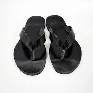May include: A pair of black leather flip-flops. The sandals have a simple design with a strap that goes between the toes and a wide strap across the top of the foot. The flip-flops are displayed on a white background.