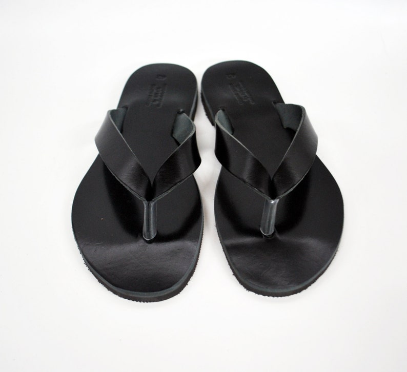 May include: A pair of black leather flip-flops. The sandals have a simple design with a strap that goes between the toes and a wide strap across the top of the foot. The flip-flops are displayed on a white background.