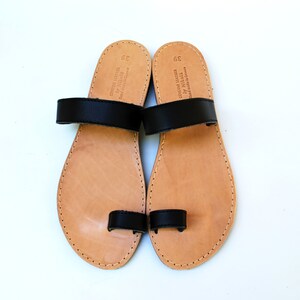 May include: A pair of handmade leather sandals with black straps. The sandals have a light brown footbed and are size 39. The text "GENUINE LEATHER by Nikola Handmade in Greece" is visible on the footbed.