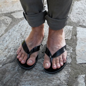 May include: Black leather flip-flops with a simple design. The sandals have a thong strap and a flat sole. The image shows the sandals on a person's feet, with rolled-up gray pants and a stone surface.