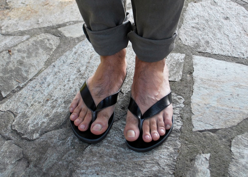 May include: Black leather flip-flops with a simple design. The sandals have a thong strap and a flat sole. The image shows the sandals on a person's feet, with rolled-up gray pants and a stone surface.