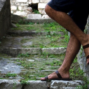 May include: Brown leather sandals with a simple strap design. The sandals are worn on a stone staircase with green vegetation. The image shows the sandals in a relaxed outdoor setting.