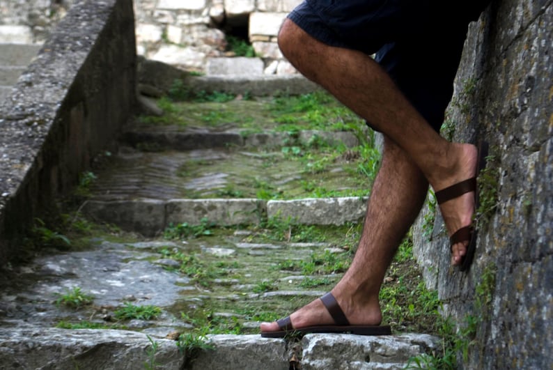 May include: Brown leather sandals with a simple strap design. The sandals are worn on a stone staircase with green vegetation. The image shows the sandals in a relaxed outdoor setting.