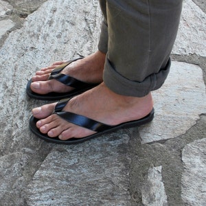 May include: Black flip-flops with a simple strap design. The sandals are worn on a person's feet, with the toes visible. The person is wearing olive green pants. The sandals are on a stone surface.