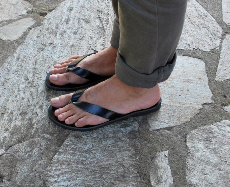 May include: Black flip-flops with a simple strap design. The sandals are worn on a person's feet, with the toes visible. The person is wearing olive green pants. The sandals are on a stone surface.