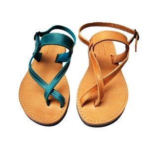 May include: A pair of leather sandals, one teal and one tan. The sandals have a toe loop and ankle strap with a buckle. The soles are light brown with visible stitching around the edges. The straps cross over the foot.