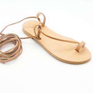 K&ouml;nnte beinhalten: Hellbraune Ledersandalen mit Zehenschlaufe und langen Kn&ouml;chelriemen. Die Sandalen haben ein minimalistisches Design, ideal f&uuml;r warmes Wetter. Die Riemen sind zur Seite gerollt.
