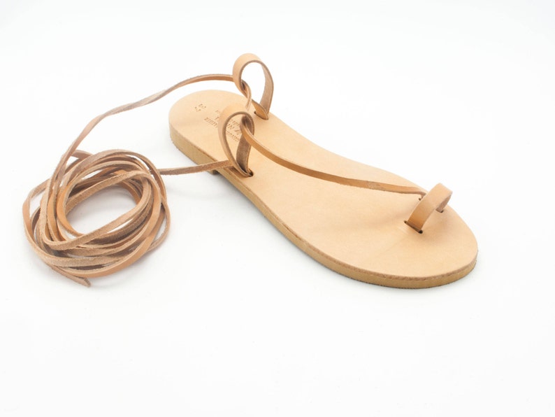 K&ouml;nnte beinhalten: Hellbraune Ledersandalen mit Zehenschlaufe und langen Kn&ouml;chelriemen. Die Sandalen haben ein minimalistisches Design, ideal f&uuml;r warmes Wetter. Die Riemen sind zur Seite gerollt.