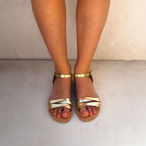 May include: Gold and tan sandals with multiple straps and ankle straps. The sandals have a flat sole and are shown from a top-down perspective. The background is a light gray color.