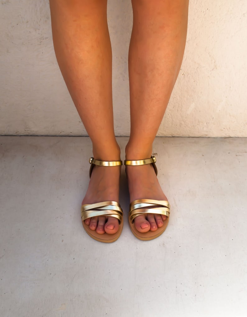 May include: Gold and tan sandals with multiple straps and ankle straps. The sandals have a flat sole and are shown from a top-down perspective. The background is a light gray color.