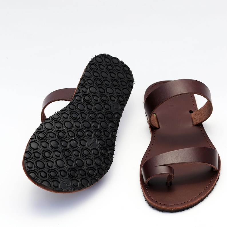 May include: A pair of brown leather sandals with a toe loop and two straps. The soles are black with a textured pattern. The sandals are displayed on a white background.