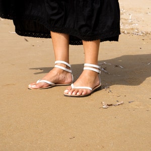 May include: White leather sandals with ankle straps and toe thong design. The sandals are worn on a sandy beach, with a black dress visible above the feet. The sandals have a minimalist design.