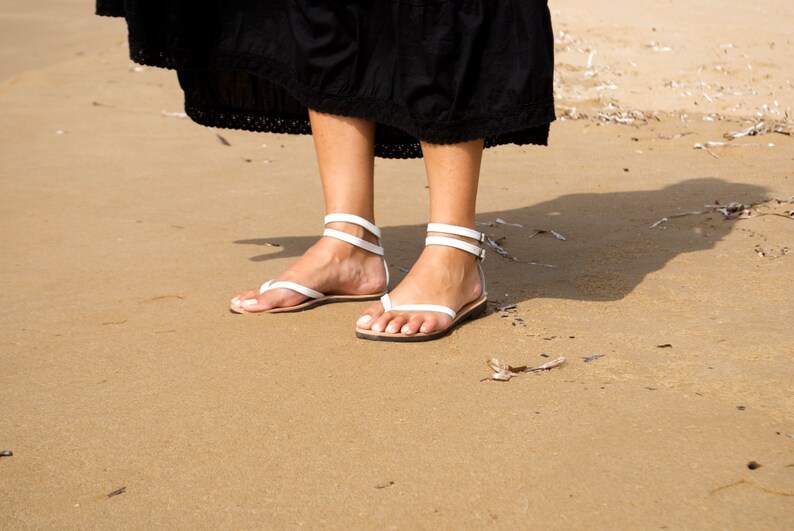 May include: White leather sandals with ankle straps and toe thong design. The sandals are worn on a sandy beach, with a black dress visible above the feet. The sandals have a minimalist design.