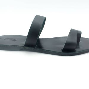 May include: Black leather sandal with two straps. The sandal has a simple design and a flat sole. The sandal is likely handmade and suitable for warm weather. The sandal is shown on a white background.