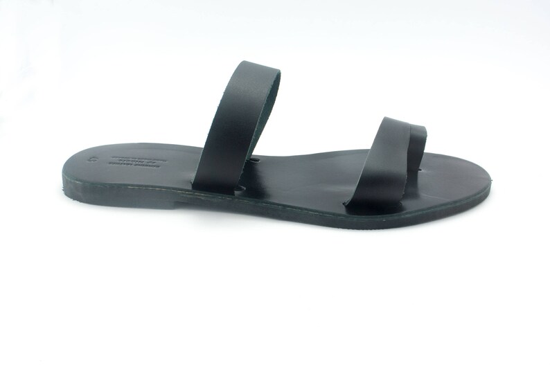 May include: Black leather sandal with two straps. The sandal has a simple design and a flat sole. The sandal is likely handmade and suitable for warm weather. The sandal is shown on a white background.