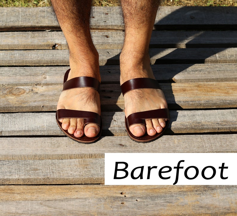 May include: Brown leather sandals with a toe loop and two straps. The sandals are shown on a wooden surface. The word "Barefoot" is written in a white rectangle below the sandals.