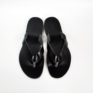 May include: A pair of black leather flip-flops, viewed from above, against a white background. The sandals have a simple design with a thong strap and a rounded toe. The leather appears smooth and shiny.