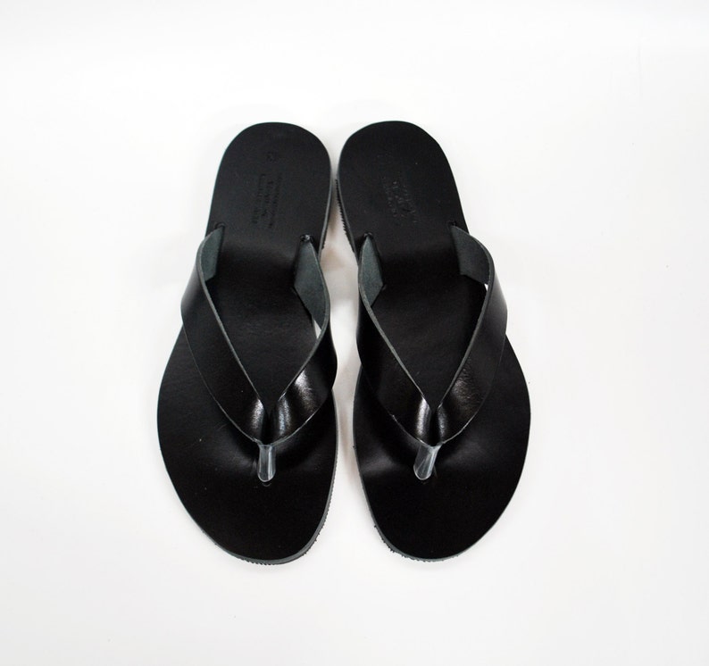 May include: A pair of black leather flip-flops, viewed from above, against a white background. The sandals have a simple design with a thong strap and a rounded toe. The leather appears smooth and shiny.