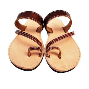 Puede incluir: Un par de sandalias de cuero marr&oacute;n con una presilla para el dedo gordo y una correa en el tobillo. Las sandalias tienen una plantilla de color beige claro y est&aacute;n cosidas con hilo blanco. Las suelas son negras.