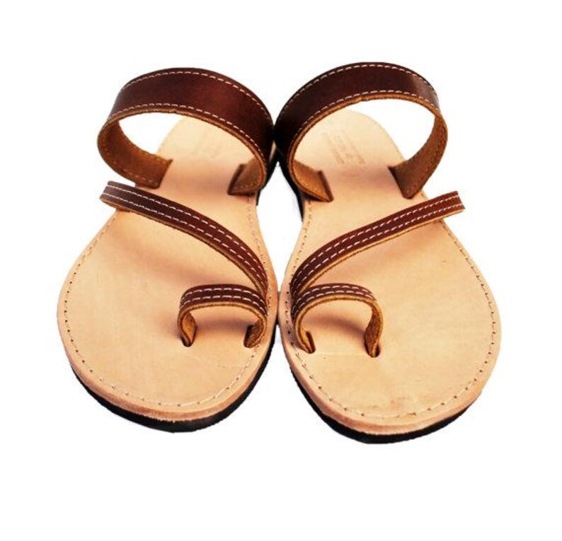 Puede incluir: Un par de sandalias de cuero marr&oacute;n con una presilla para el dedo gordo y una correa en el tobillo. Las sandalias tienen una plantilla de color beige claro y est&aacute;n cosidas con hilo blanco. Las suelas son negras.