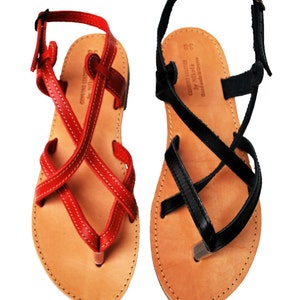 May include: A pair of leather sandals, one red and one black, with crisscross straps and toe loops. The sandals have a natural leather footbed and adjustable ankle straps. The red sandal has white stitching, and the black sandal has black stitching. The sandals are labeled "Genuine Leather by Nikota, Hand made in Greece."