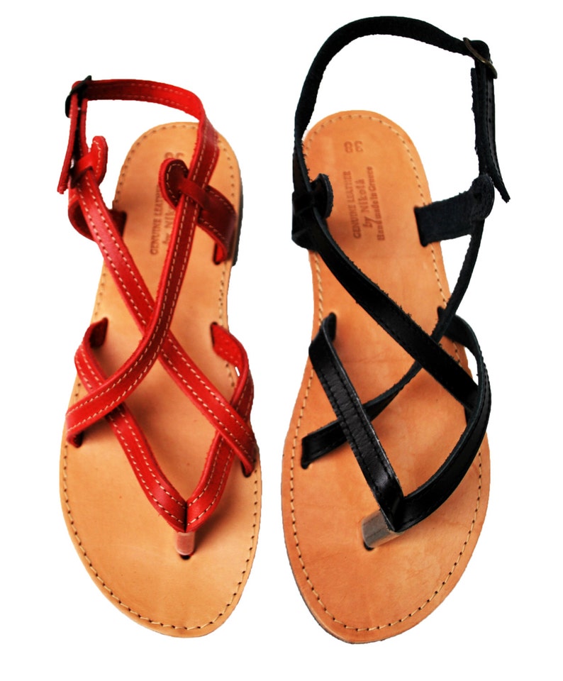 May include: A pair of leather sandals, one red and one black, with crisscross straps and toe loops. The sandals have a natural leather footbed and adjustable ankle straps. The red sandal has white stitching, and the black sandal has black stitching. The sandals are labeled "Genuine Leather by Nikota, Hand made in Greece."