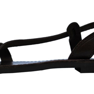May include: A dark brown leather sandal with a flat sole. The sandal features a toe strap, an ankle strap with a buckle, and a strap that goes around the ankle. The sandal is designed for comfort and style.