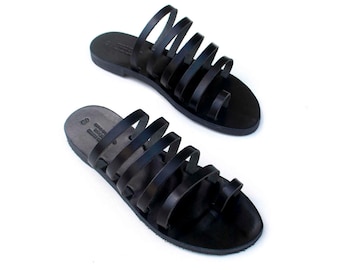 May include: A pair of black leather sandals with multiple straps. The sandals have a toe loop and a flat sole. The footwear is displayed on a white background.