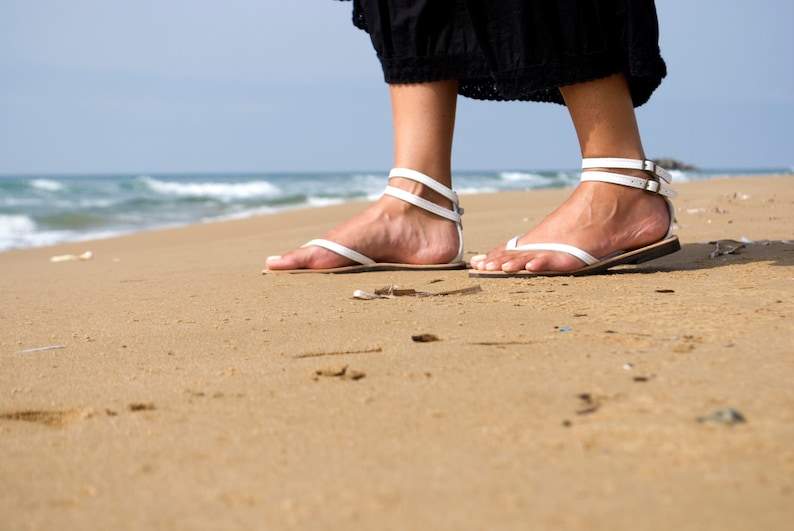 May include: White leather sandals with ankle straps and toe loops. The sandals are worn on a sandy beach with the ocean in the background. The straps have silver-colored buckles. The sandals have a flat sole.