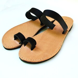 May include: A pair of leather sandals with a toe loop and a strap across the top of the foot. The sandals have a light brown footbed and black straps. The edges are stitched with a light-colored thread.