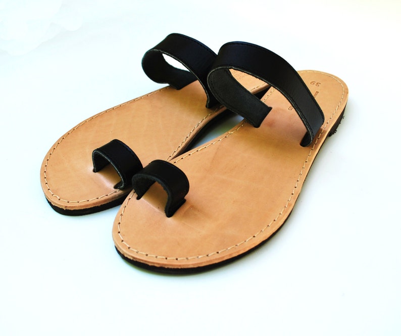 May include: A pair of leather sandals with a toe loop and a strap across the top of the foot. The sandals have a light brown footbed and black straps. The edges are stitched with a light-colored thread.